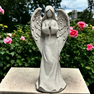 May include: A gray concrete angel statue with wings outstretched and hands clasped in prayer. The statue is standing on a gray concrete base. The statue is surrounded by pink roses.