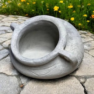 May include: A gray concrete planter with a textured surface and a rounded shape. The planter has a wide opening at the top and a smaller opening on the side.