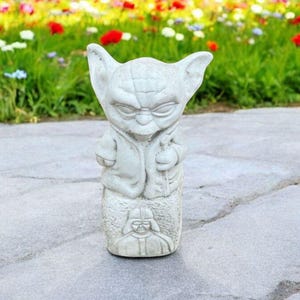 以下が含まれることがあります： スターウォーズのヨーダの白いセラミック像。ヨーダは茶色のローブを着ており、顔には真剣な表情をしています。彼はダース・ベイダーを描いた台座の上に立っています。