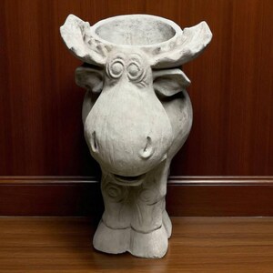 May include: A light gray moose-shaped planter. The planter has a large head with prominent antlers and a wide, open mouth. The body of the moose is rounded, and the legs are short and sturdy. The planter is made of a textured material and is suitable for indoor or outdoor use.