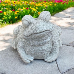 May include: A gray concrete frog statue with large eyes and a textured surface. The frog is sitting on a stone path, with a vibrant flower garden in the background.