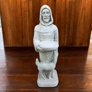 May include: A gray stone statue of a robed figure with a hood, holding a shallow bowl. A small deer stands at the figure's feet. The statue is set against a wooden background.