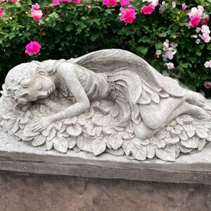 May include: A concrete sculpture of a sleeping fairy with wings, lying on a bed of leaves. The sculpture is on a gray stone base.