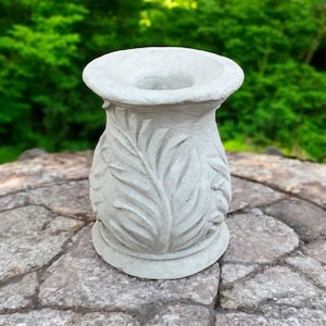 May include: A gray concrete vase with a leaf design. The vase is sitting on a stone surface.