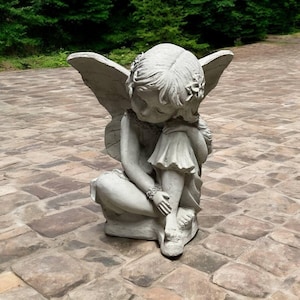 May include: A gray stone sculpture of a fairy with wings, sitting with her head resting on her hand. The fairy is wearing a dress and has flowers in her hair.