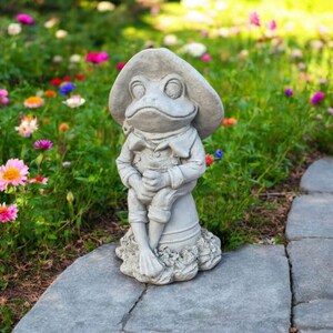 May include: A gray concrete frog statue wearing a hat and overalls, seated on a pedestal. The frog is holding its legs and has a whimsical expression. The statue is placed on a stone path in a garden with colorful flowers.