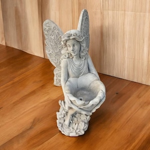 Puede incluir: Estatua de hada de hormigón gris con alas detalladas y una corona floral. El hada sostiene un cuenco grande en forma de concha, probablemente para un baño de pájaros o uso decorativo. La estatua está sobre un fondo de madera.