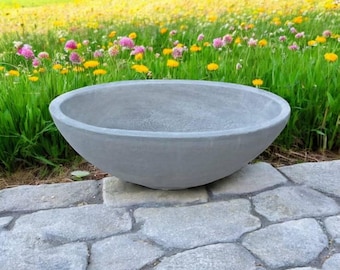 May include: A large, gray, concrete planter bowl with a rounded shape. The bowl sits on a stone patio with a backdrop of a vibrant green lawn filled with wildflowers. The planter is designed for outdoor use.