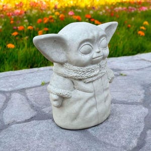 Pu&ograve; includere: Una statua in cemento grigio di un personaggio del franchise di Star Wars, che indossa una giacca con cappuccio e una sciarpa. La statua &egrave; in piedi su una superficie di pietra grigia.