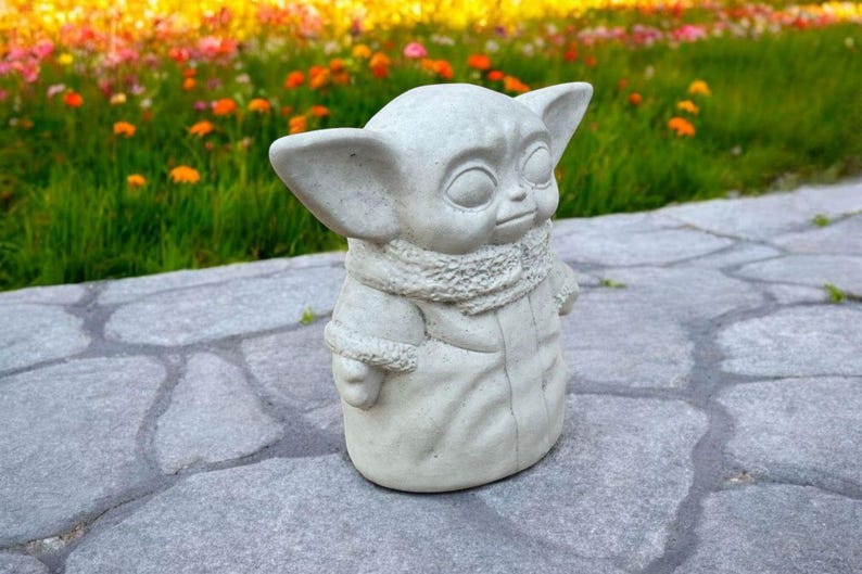 Pu&ograve; includere: Una statua in cemento grigio di un personaggio del franchise di Star Wars, che indossa una giacca con cappuccio e una sciarpa. La statua &egrave; in piedi su una superficie di pietra grigia.