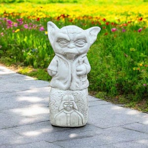 以下が含まれることがあります： スターウォーズのヨーダの白いコンクリート製のガーデン像で、ダース・ベイダーを描いた台座の上に立っています。像は庭の石畳の上にあります。