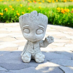May include: A gray concrete statue of a character from the Guardians of the Galaxy movie. The statue is in a sitting position with one arm raised and a friendly expression on its face.