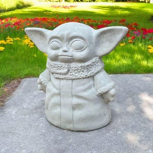 Pu&ograve; includere: Una statua di cemento grigio di un personaggio immaginario del franchise di Star Wars. Il personaggio indossa una tunica con cappuccio e ha grandi orecchie.