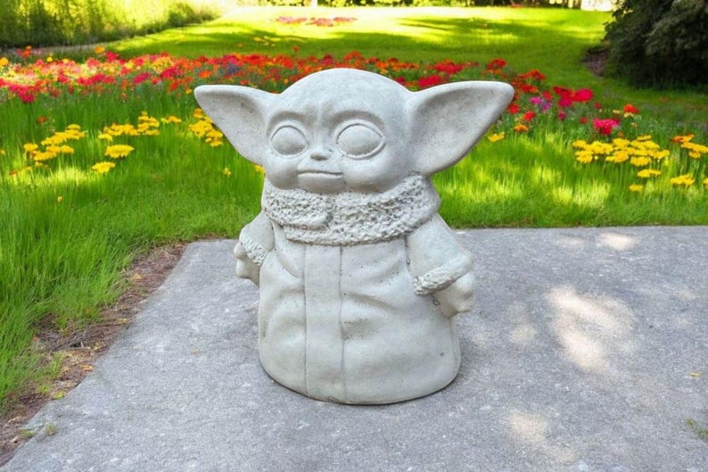 Pu&ograve; includere: Una statua di cemento grigio di un personaggio immaginario del franchise di Star Wars. Il personaggio indossa una tunica con cappuccio e ha grandi orecchie.