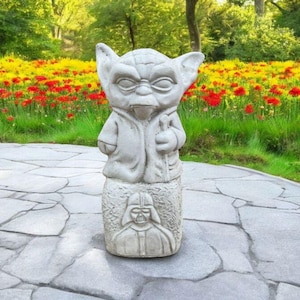 以下が含まれることがあります： 石畳の上に立っている、スターウォーズのヨーダのグレーのコンクリート像。ヨーダは特徴的な茶色のローブを着ており、ライトセーバーを持っています。像は赤と黄色の花壇に囲まれています。