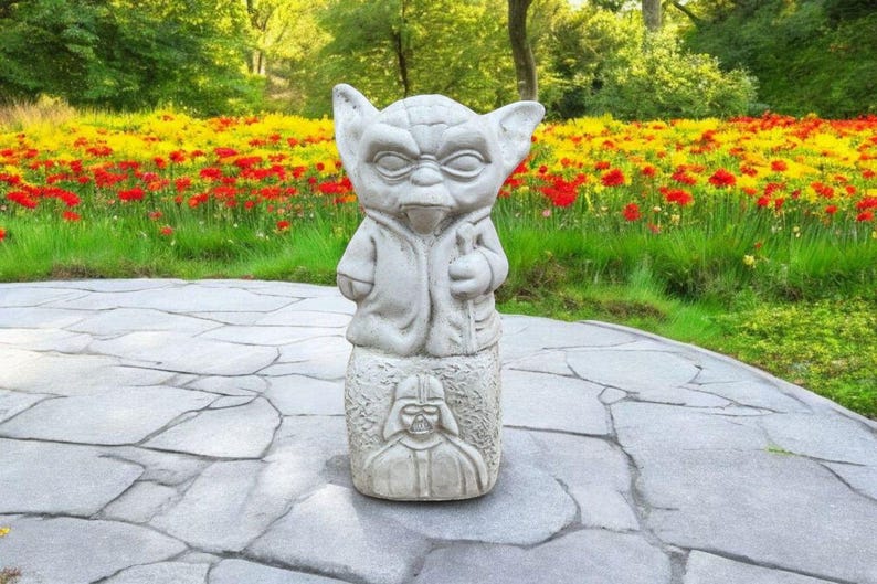 以下が含まれることがあります： 石畳の上に立っている、スターウォーズのヨーダのグレーのコンクリート像。ヨーダは特徴的な茶色のローブを着ており、ライトセーバーを持っています。像は赤と黄色の花壇に囲まれています。