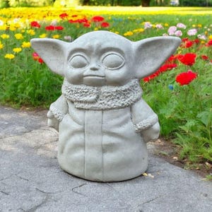 Pu&ograve; includere: Una statua di cemento grigio di un personaggio immaginario con grandi orecchie e una tunica con cappuccio. La statua &egrave; in piedi su un sentiero di cemento grigio in un giardino con fiori.