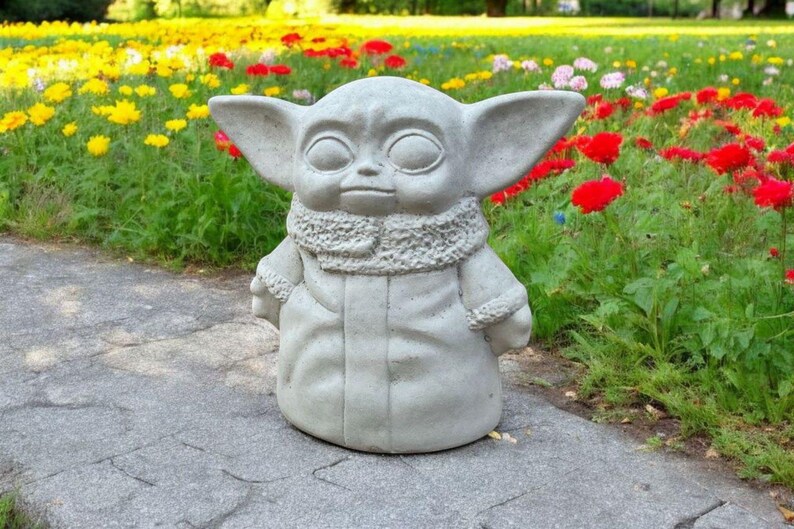 Pu&ograve; includere: Una statua di cemento grigio di un personaggio immaginario con grandi orecchie e una tunica con cappuccio. La statua &egrave; in piedi su un sentiero di cemento grigio in un giardino con fiori.