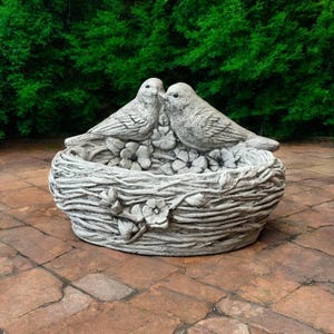 May include: A concrete bird bath shaped like a bird's nest with two birds perched on the edge. The nest is decorated with flowers and vines.