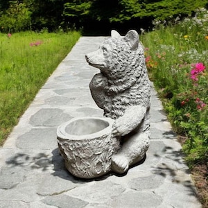 May include: A gray concrete bear statue holding a planter pot. The bear is sitting on a stone path in a garden setting. The planter pot is shaped like a tree stump.