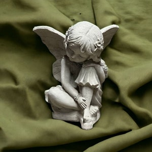May include: A white, ceramic figurine of a fairy with wings, sitting with her head down and hands clasped together. The fairy is wearing a dress with a ruffled hem.