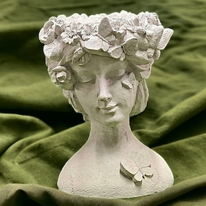 May include: A white ceramic planter shaped like a woman's bust with a floral crown and a butterfly on the chest. The planter is on a green fabric background.