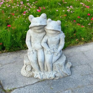 May include: A pair of gray concrete frog statues sitting on a tree stump. The frogs are facing each other and appear to be holding hands.