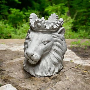 May include: A gray concrete lion head planter with a crown on top. The planter is sitting on a stone surface.