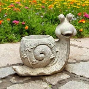 May include: A gray concrete snail planter with a detailed shell and a whimsical face. The planter is sitting on a stone path.