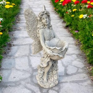 May include: A stone fairy statue with detailed wings and a floral basket. The fairy is dressed in a flowing gown and stands on a base of flowers. The statue is displayed on a stone path with flowers in the background.