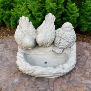 May include: A gray concrete bird bath featuring three detailed bird sculptures perched above a basin. The birds are facing downward, with textured feathers. The bird bath is set on a stone surface, with green foliage in the background.