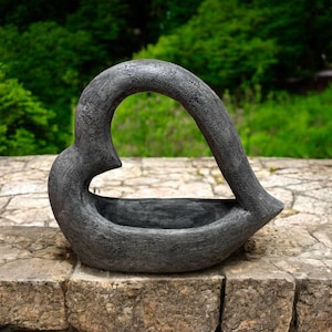 May include: A black heart-shaped planter made of concrete. The planter has a rough, textured surface and a wide opening at the top.