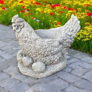 May include: A detailed, gray concrete hen planter with a textured surface. The hen is sculpted with feathers and sits atop a base with three eggs. The planter is designed to hold plants and is suitable for outdoor use.