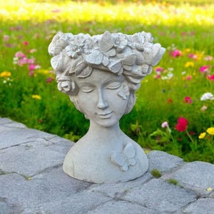 May include: A white concrete planter shaped like a woman's bust with a floral crown and butterfly accents. The planter is sitting on a gray stone path.