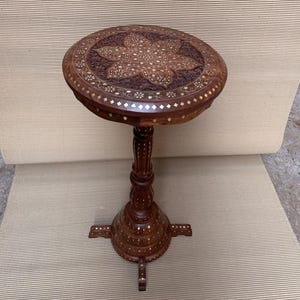May include: A small, round, dark brown wooden side table with intricate floral carvings on the tabletop. The table has a single pedestal base with four feet and decorative inlaid accents. The table is approximately 61 cm tall.