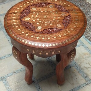 May include: A round, wooden side table with a dark brown finish. The tabletop features intricate floral carvings and inlaid gold-coloured accents. The table has three curved legs, also with gold accents, and is suitable for decorative use.