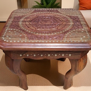 May include: A wooden coffee table with detailed floral carvings on the surface. The table is rectangular with curved legs and decorative inlays. The wood has a warm, reddish-brown colour. The table is placed on a light-coloured rug. The table measures approximately 90 cm in length.