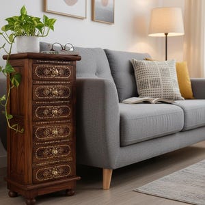 Puede incluir: Un mueble de madera oscura con seis cajones, adornado con detalles dorados. El mueble está colocado junto a un sofá gris, con una planta encima. La habitación está decorada con una lámpara y un cojín estampado.