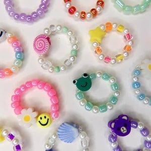 May include: A collection of colorful beaded rings with various charms. The rings feature a variety of colors, including pink, purple, blue, and yellow. Some rings have smiley faces, flowers, stars, and other decorative elements.