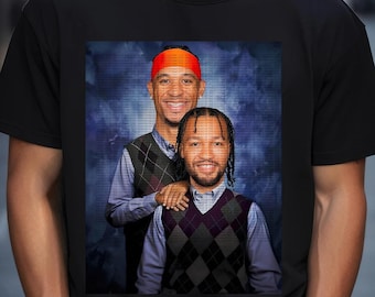 Op de afbeelding: Een zwart T-shirt met een grafische print van twee mannen in truien. De man links draagt ​​een rode hoofdband en een blauwe trui met een wit patroon. De man rechts draagt ​​een grijze trui met een zwart-wit ruitpatroon.