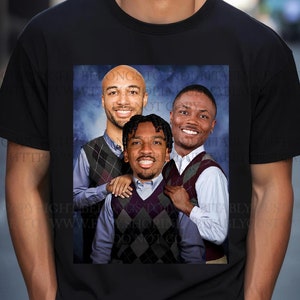 May include: A black t-shirt featuring a graphic print of three men wearing sweaters. The men are smiling and have their arms around each other. The background is a blue and white gradient.