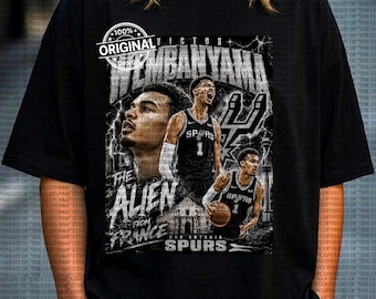 Op de afbeelding: Zwart T-shirt met een afbeelding van Victor Wembanyama in een San Antonio Spurs-shirt. Het ontwerp bevat de tekst "100% Original" en "The Alien From France". Het shirt heeft een vintage uitstraling.