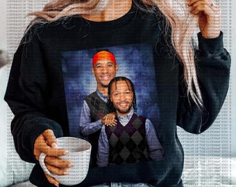 Op de afbeelding: Zwart sweatshirt met een ingelijst portret van twee mannen. Het portret toont de mannen in argyle vesten en overhemden met knopen. Eén man draagt een rode hoofdband. Het sweatshirt wordt gedragen door een persoon die een witte mok vasthoudt.