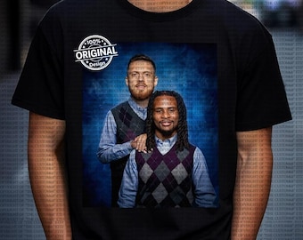 Op de afbeelding: Zwart T-shirt met een portret van twee mannen in argyle vesten en overhemden met kraag tegen een blauwe achtergrond. Het shirt heeft ook een wit cirkelvormig ontwerp met de woorden "100% Original Design".