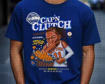 Op de afbeelding: Blauw T-shirt met een parodieontwerp van een ontbijtgranendoos. Het ontwerp bevat de tekst "CAP'N CLUTCH" en een illustratie van een basketballer. De ontbijtgranendoos leest "NATURALLY CLUTCH OAT BRUN CEREAL" en "NET WT 45.8g".