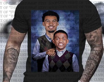 Op de afbeelding: Een zwart T-shirt met een bedrukte afbeelding van twee mannen die truien dragen. De mannen lachen en staan ​​naast elkaar. De achtergrond is een blauwe en witte gradiënt.