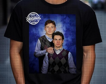 Op de afbeelding: Zwart T-shirt met een afgedrukte afbeelding van twee mannen in argyle vesten en overhemden met kraag tegen een blauwe achtergrond. Het shirt heeft ook een wit cirkelvormig ontwerp met de tekst "100% ORIGINAL Design."