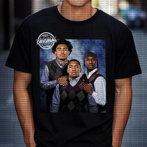 May include: Black t-shirt featuring a graphic of three men wearing argyle vests over button-down shirts. The image has a blue background. The shirt also has a circular "100% Original Design" logo.