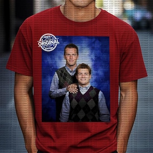 May include: A red t-shirt with a printed image of two men wearing argyle vests and collared shirts against a blue background. The shirt also has a circular "100% Original Design" graphic.