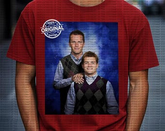 Op de afbeelding: Rood t-shirt met een bedrukte afbeelding van twee mannen in ruitjesvesten en overhemden met kraag tegen een blauwe achtergrond. Het shirt heeft ook een cirkelvormige "100% Original Design" afbeelding.